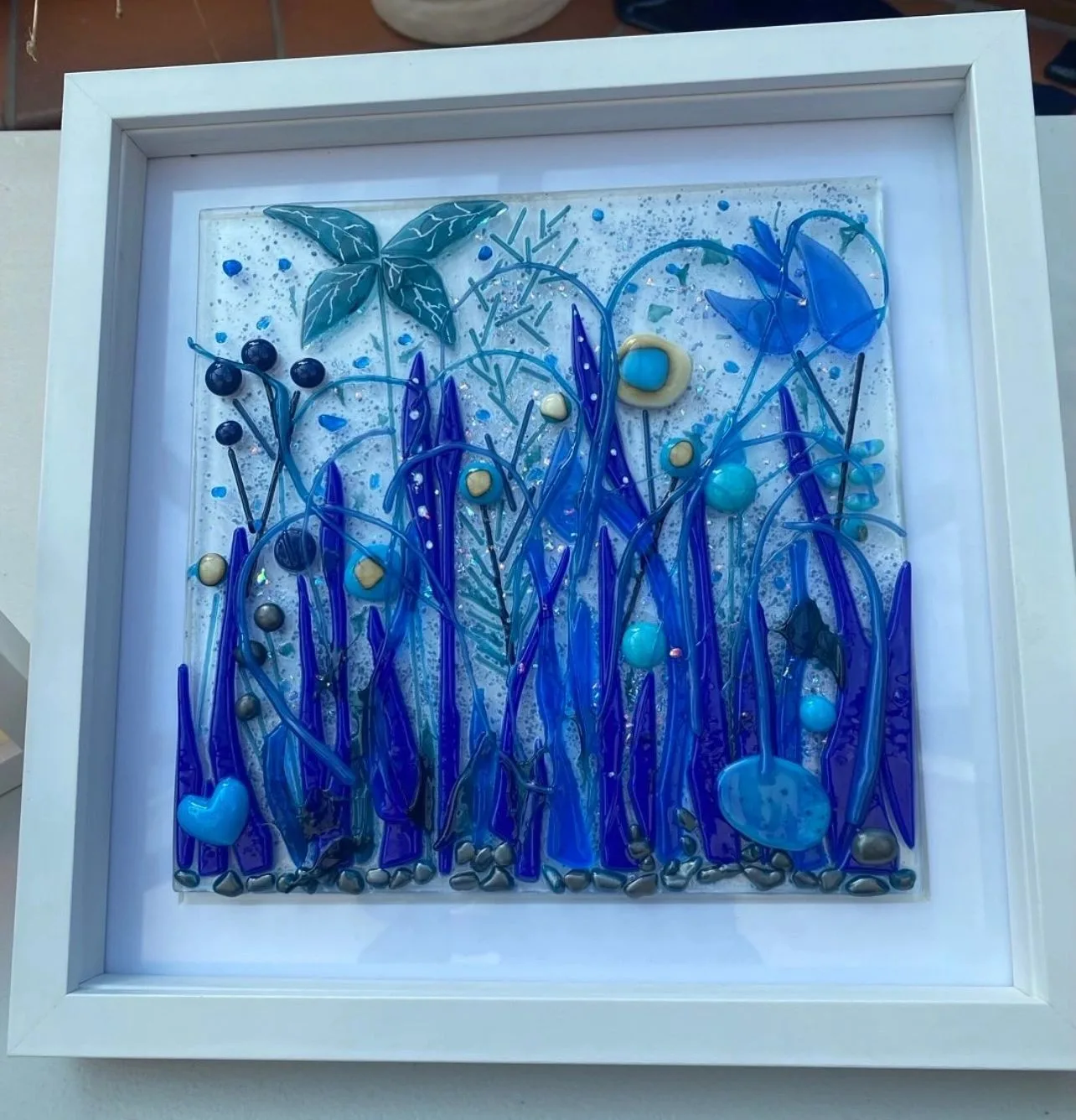 A framed fused glass picture of blue and purple flowers and grasses